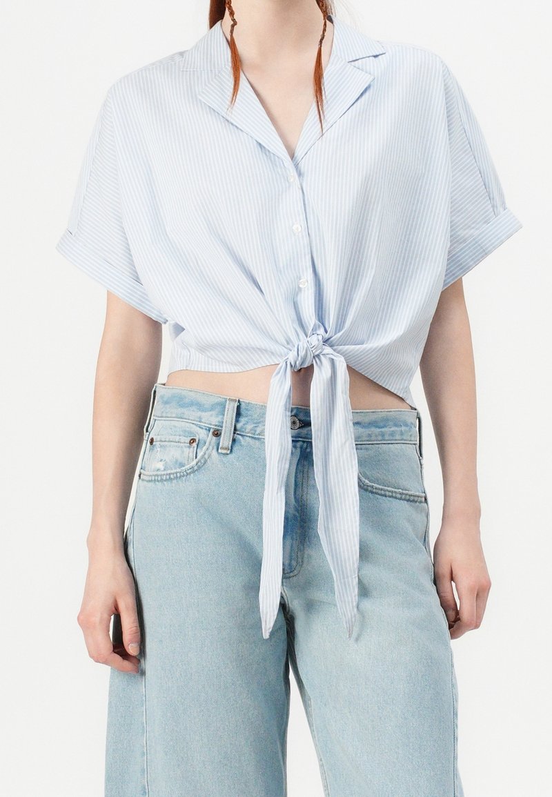 Chemise à manches courtes rayée bleu et blanc avec un nœud à la taille, associée à un jean en denim bleu clair taille haute. Texture de tissu lisse.