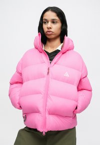 Chaqueta de plumón rosa con cuello alto, cierre de cremallera y costuras visibles. Características notables incluyen un logo y dobladillo ajustable.