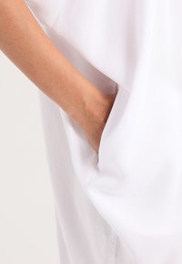 Indumento bianco con una vestibilità ampia e una tasca laterale, realizzato in tessuto morbido e con una consistenza liscia. La manica si adagia delicatamente sul braccio.