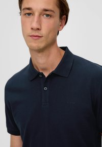 Tamno mornarica polo majica od teksturiranog materijala, s klasičnom kragnom, plavim plaketom s dva dugmeta i malim izvezenim logom na prsima.