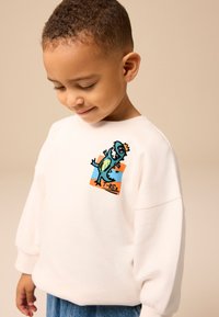 Hanorac alb cu grafică T-Rex în culorile teal, portocaliu și negru. Are o croială lejeră și manșete cu nervuri, asortat cu pantaloni din denim.