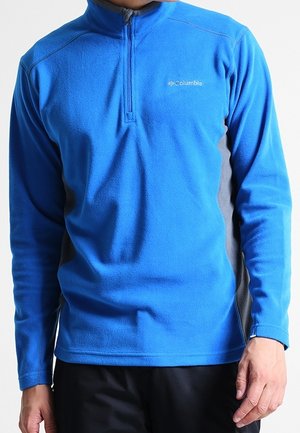 Homme portant un pull polaire bleu Columbia avec une fermeture éclair à moitié et des panneaux latéraux gris, debout, les bras détendus le long du corps.