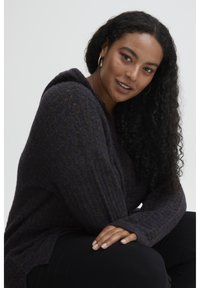 Pull en tricot sombre avec capuche, présentant un tissu texturé et des manches côtelées. Le mannequin a les cheveux bouclés et est assis contre un fond neutre.