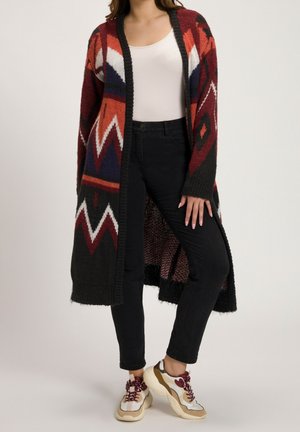 Kvinde iført en lang, mønstret cardigan med røde, sorte, hvide og marineblå zigzag-designs over en hvid top og sorte jeans med beige og mørkerøde sneakers.