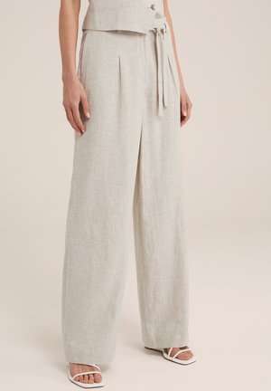 Mujer con pantalones amplios de color beige claro con pliegues frontales y cinturón atado, combinados con sandalias blancas de tacón abierto.
