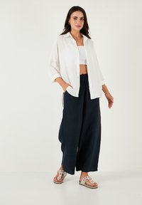 White, button-up shirt with a textured fabric, worn over a white crop top, paired with navy wide-leg trousers and decorative sandals.