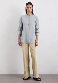 Chemise bleu clair à boutons, à manches longues, en tissu doux. Associée à un pantalon beige ample et des sandales noires.