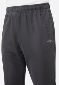 Nahaufnahme von schwarzen Sporthosen mit elastischem Bund, die eine Reißverschlusstasche an der Seite und das Logo "SPORT by STOCKER" auf dem rechten Oberschenkel aufweisen.