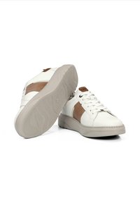 Zapatillas blancas con acentos de tela marrón, con una suela de goma gruesa, puntera redonda y cordones planos. Detalles texturizados en los laterales.