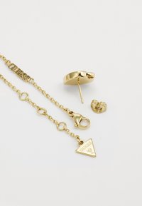 Ensemble de bijoux en ton or comprenant des boucles d'oreilles rondes à clou, une chaîne avec un fermoir à homard et un pendentif en forme de triangle gravé avec "GUESS."