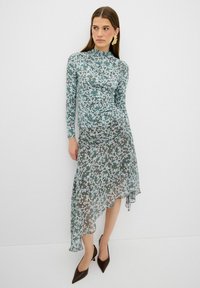 Langärmliges Blumenkleid in hellblauen, grünen und rosafarbenen Farbtönen; asymmetrischer Saum; durchscheinender Stoff; kombiniert mit braunen spitzen Absätzen.