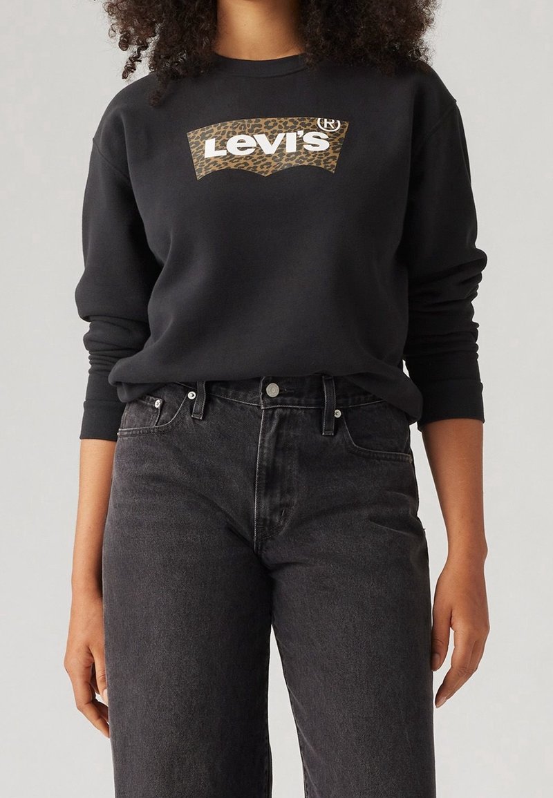 Femme portant un sweat-shirt noir Levi's avec un logo imprimé léopard et un jean noir délavé taille haute sur un fond clair uni.
