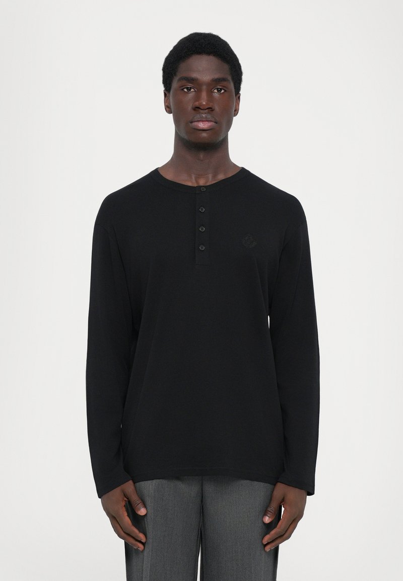 Čierna dlhá rukávová henley košeľa s okrúhlym výstrihom, zapínaním na štyri gombíky a jemným logo detailom na hrudi. Hladká tkanina a voľný strih.