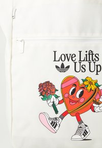 Canvas tote bag in cream featuring a cartoon heart character holding roses. Printed text "Love Lifts Us Up" and black Adidas logo.