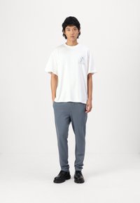 Camiseta blanca de algodón con un estampado gráfico, combinada con pantalones grises holgados y zapatos negros. Fondo neutro, pose de pie.