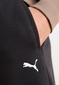 Czarne spodnie sportowe mają boczną kieszeń, a na udzie znajduje się białe logo Pumy. Materiał wydaje się gładki i elastyczny.