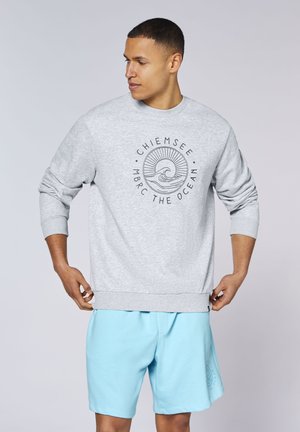 Man wearing light gray sweatshirt with ocean wave logo and text "Chiemsee MBRC The Ocean" and light blue shorts looking left.