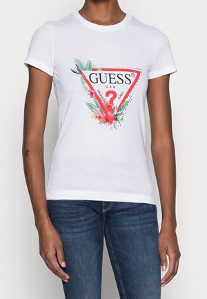 Person iført en hvid t-shirt med korte ærmer med et rødt trekantet Guess-logo, blomsterdekorationer og blå jeans.