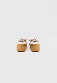 Pari mokkasinslippers i beige semsket skinn med hvit pelsfôr og sløyfedetaljer, vist fra baksiden på en hvit bakgrunn.