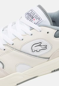 Sněrky Lacoste s bílou kůží a šedými akcenty, kulaté tkaničky, síťovinou a texturovaným logem krokodýla na straně.