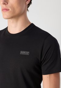 Tricou din bumbac negru, cu guler rotund și mâneci scurte, având o mică eticheta cu logo gri pe partea stângă a pieptului.