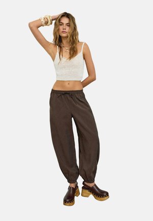 Frau trägt ein weißes gehäkeltes Kurztop, braune weite Hosen, braune Holz-Plateauschlappen und mehrere Armbänder, steht mit erhobenem Arm.