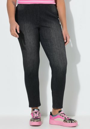 Henkilö, joka pukeutuu mustiin istuviin farkkuihin, kirkkain pinkkeihin ja leopardikuvioisiin tennareihin, seisoo valkoista seinää vasten kädet rentoina sivuilla.