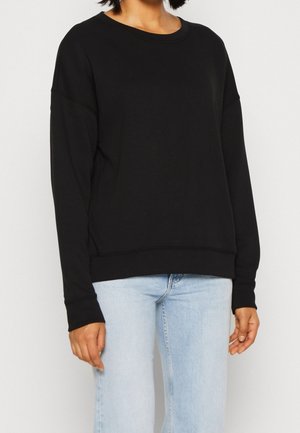 Person iført en ensfarvet sort langærmet sweatshirt og lyseblå jeans, stående mod en ensfarvet hvid baggrund.