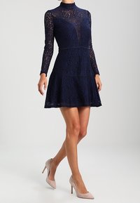 Marineblaues Spitzenkleid mit langen Ärmeln und hohem Ausschnitt, ausgestelltem Rock, Blumenmustern und subtiler Textur. Kombiniert mit hellrosafarbenen Absätzen.