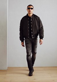 Chaqueta bomber negra con botones a presión, camiseta negra, jeans grises desgastados y gafas de sol negras. El modelo camina sobre un suelo de madera frente a una pared lisa.