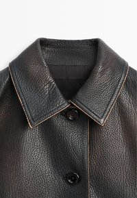 Colletto di giacca in pelle nera con cuciture marroni visibili e due bottoni neri su tessuto di pelle lavorata.