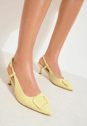 Woman wearing pale yellow woven slingback kitten heels with large square buckle on toes, standing on light floor.
