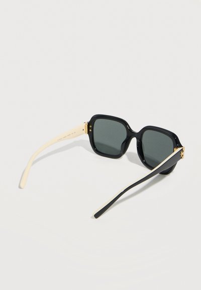 Lunettes de soleil à monture carrée noire avec des verres foncés et des branches beige contrastantes, posées en angle sur une surface blanche.