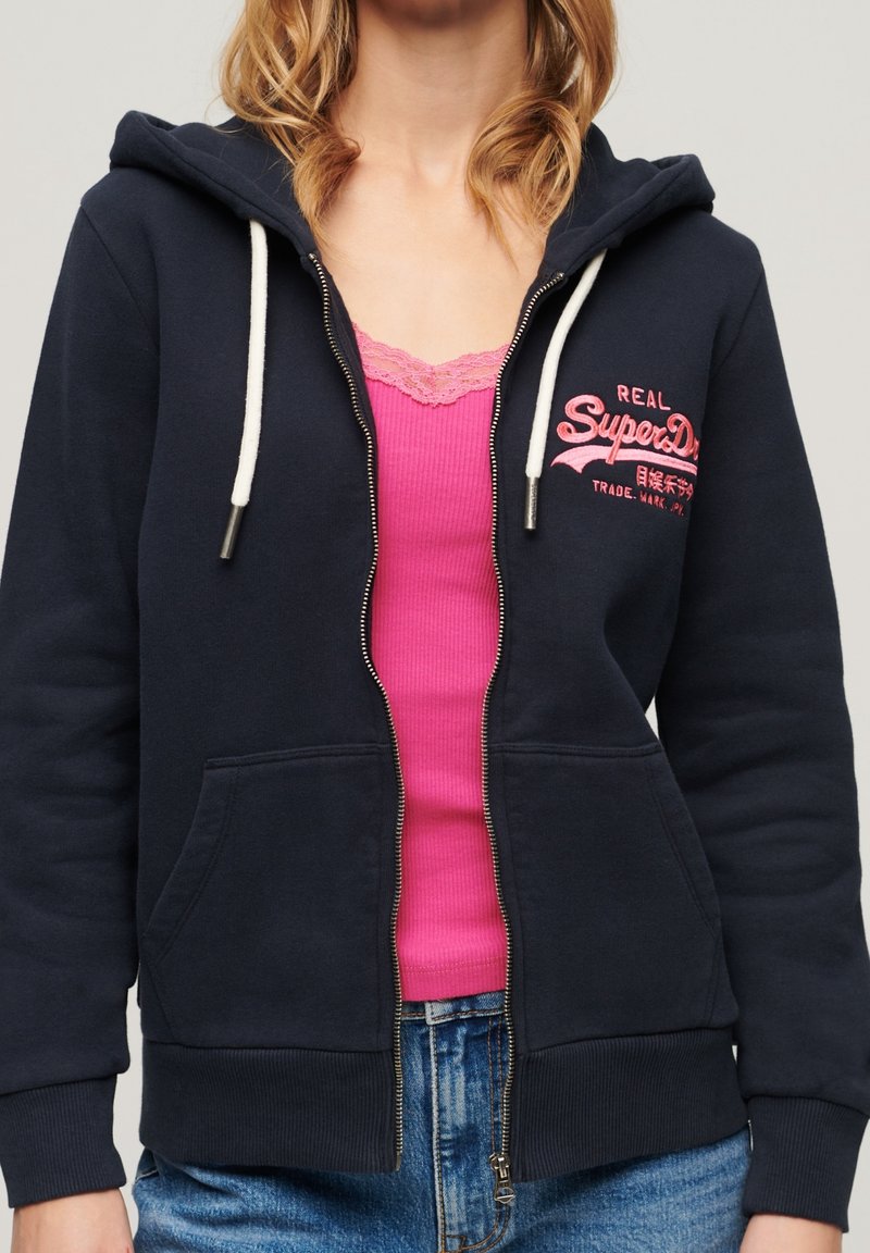 Felpa con zip blu navy, con lacci bianchi, tasche a marsupio e testo ricamato rosa. Indossata sopra una canotta rosa a coste e jeans in denim.