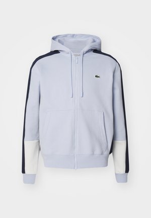 Jasnoniebieska bluza z kapturem na suwak z granatowymi paskami na rękawach, białymi mankietami, przednimi kieszeniami, kapturem ze sznurkami oraz małym zielonym logo krokodyla na piersi.