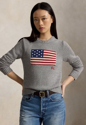 Femme aux longs cheveux foncés portant un pull gris avec un motif du drapeau américain et un jean bleu, les mains sur les hanches.