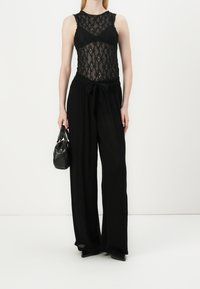 Body de encaje negro con un patrón intrincado, combinado con pantalones negros holgados que cuentan con una cinturilla anudada. La modelo sostiene un pequeño bolso negro.