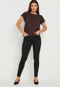 Top corto marrone con scollo rotondo e dettagli laterali, abbinato a jeans skinny neri e scarpe con tacco nere.