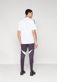 Camisa blanca de manga corta, pantalones deportivos morados con patrones blancos y rayas laterales, llevados con zapatos negros; vistos desde atrás.