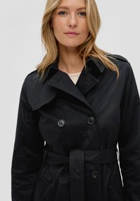 Manteau noir en trench avec un design à double boutonnage, grand col, boutons assortis et une ceinture en tissu nouée autour de la taille. Texture lisse.