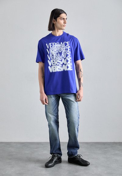 T-shirt en coton bleu avec un design graphique blanc comprenant un texte répété et un motif central de visage, associé à un jeans bleu clair et des chaussures noires.