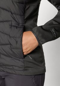 Veste noire isolée présentant une texture matelassée d'un côté, avec une finition lisse de l'autre, poche zippée et poignets ajustables.