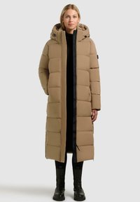 Lange, puffer jas in tan met een capuchon, voorzien van een ritssluiting aan de voorkant, zwarte accenten en een horizontaal gestikt patroon. Geschikt voor koud weer.