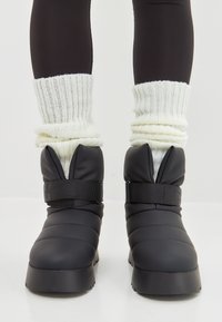 Botas de plumas negras con un diseño acolchado, que presentan una amplia correa de Velcro y punta redondeada. Combinadas con calcetines de punto blanco con acanalado.