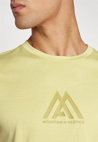Šviesiai geltona marškinėlių su tekstūruotu audiniu, kuriuose yra ryškus iškilus logotipas su kalno grafika ir užrašu "MOUNTAIN ATHLETICS."