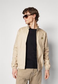 Beige lett jakke med ståkrage, frontglidelås og elastiske mansjetter. Svart strikkegenser under, parret med mørke solbriller.