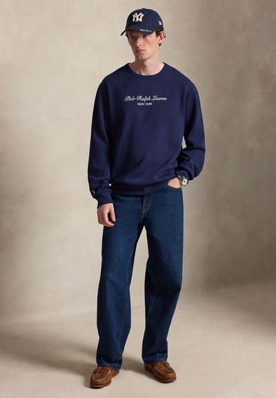 Άνδρας που φοράει μπλε σκούρο φούτερ Polo Ralph Lauren, σκούρο μπλε τζιν, καφέ παπούτσια και μπλε σκουφάκι μπέιζμπολ New York Yankees, στέκεται μπροστά σε μπεζ φόντο.