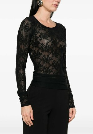 Donna che indossa un top a maniche lunghe in pizzo floreale nero con pantaloni neri e grandi orecchini rotondi dorati, in piedi davanti a uno sfondo bianco.