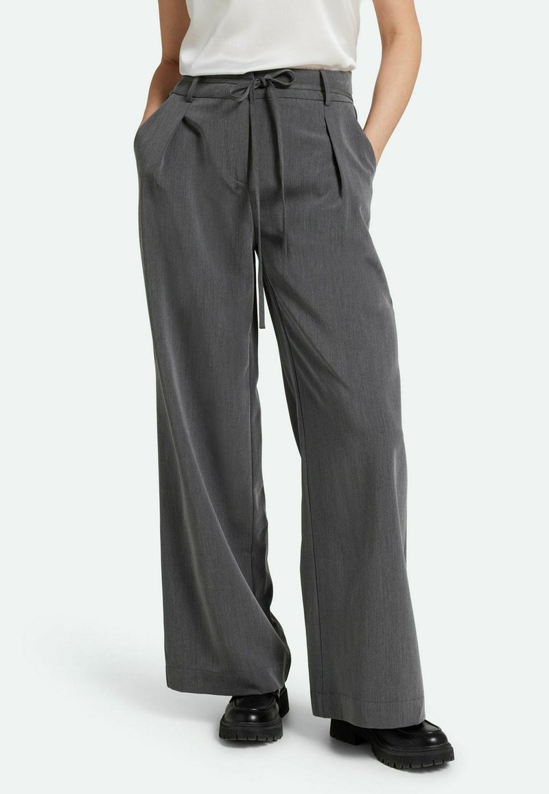 Pantalones de pierna ancha grises con cintura de cordón, con pliegues y bolsillos laterales. Están hechos de una tela texturizada y combinados con zapatos de plataforma negros.