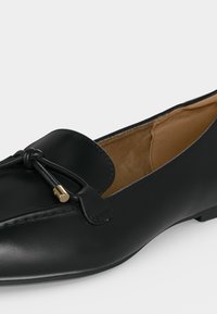 Mocassin en cuir noir avec bout arrondi, nœud décoratif et embouts métalliques dorés, avec une doublure intérieure beige.